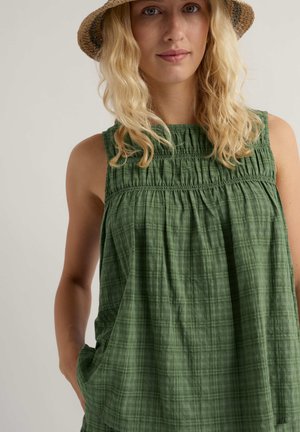 Blondinka z vpletenim rjavim klobukom in zeleno obleko brez rokavov z reliefnim vzorcem in obročem na prsih, stoji z eno roko v žepu na enobarvnem ozadju.