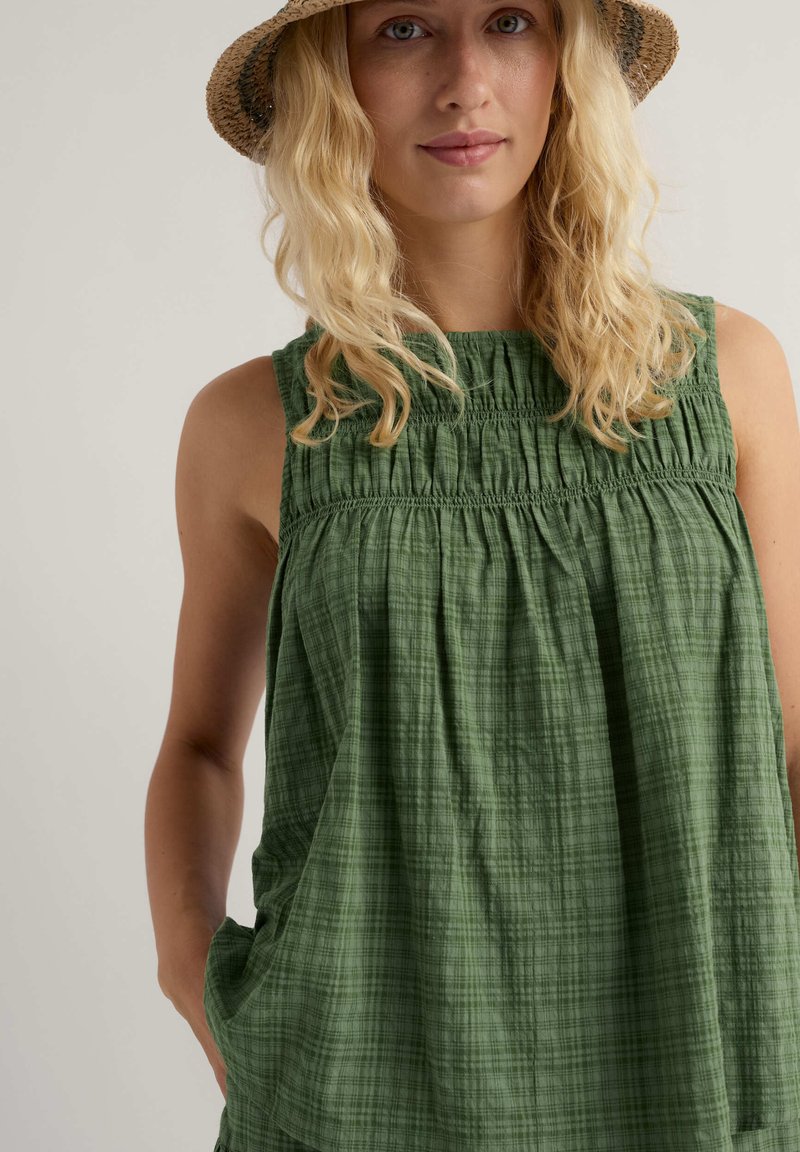 Blonde Frau mit einem gewebten, hellbraunen Hut und einem ärmellosen, grünen, strukturierten Kleid mit gerafftem Bruststück, die mit einer Hand in der Tasche vor einem schlichten Hintergrund steht.