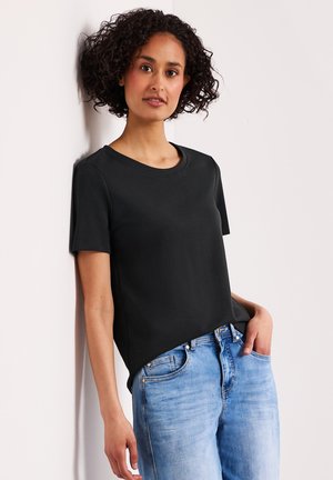 Kobieta z kręconymi włosami opierająca się o białą ścianę, ubrana w czarną koszulkę z krótkim rękawem i jasnoniebieskie dżinsy, z jedną ręką w kieszeni.