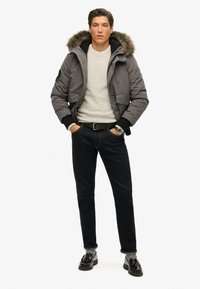 Giubbotto puffer grigio con cappuccio foderato in pelliccia, abbinato a un leggero maglione, jeans in denim scuro e mocassini neri. Include un orologio da polso in argento.