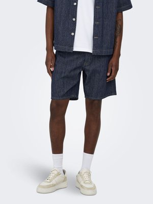 Homme portant un short en jean bleu foncé, une chemise assortie à manches courtes avec boutons, des chaussettes blanches montantes et des baskets écrues, debout devant un fond uni.