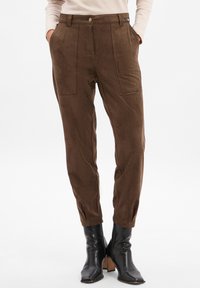 Bruine hoge taille broeken met opgestikte zakken, gedragen met zwarte hakken enkellaarsjes en een beige langemouwtop.