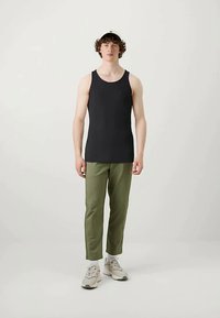 Fiatal férfi áll, fekete ujjatlan bordázott felsőt, olíva zöld nadrágot, fehér zoknit és bézs sportcipőt visel egy sima háttér előtt.