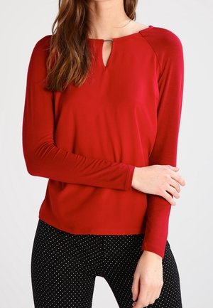 Nainen, jolla on pitkähihaisen punaisen paidan etuosassa pieni avonainen yksityiskohta ja mustat housut, joissa on valkoisia pilkkuja, seisoo kädet ristissä.