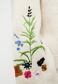 Pull en maille couleur crème orné d'une broderie florale vibrante en rose, bleu, noir et orange. Texture dense avec des coutures détaillées.