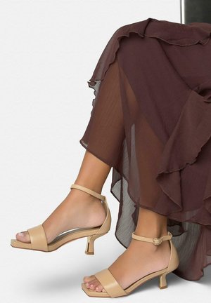 Pieds portant des sandales à talons avec bride cheville beige, associées à une jupe transparente marron à volants, jambes croisées sur un fond uni.