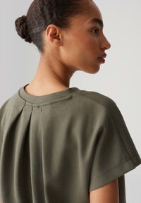 Olive green top with short sleeves, featuring a pleated back detail and smooth texture. Clean lines and minimal design.
