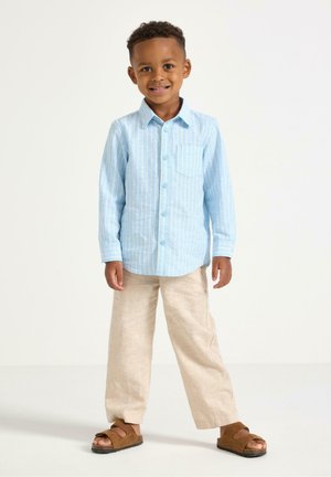 Ung dreng smilende, iført en lyseblå, stribet langærmet skjorte, beige bukser og brune sandaler, stående mod en ensfarvet baggrund.