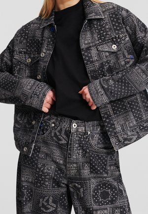 Vrouw draagt een zwarte en grijs gemengde spijkerjas en bijpassende broek met een zwart shirt eronder, handen die de voorkant van de jas vasthouden.