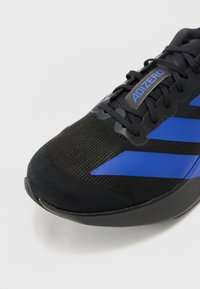 Chaussure de sport noire avec une tige texturée et des accents bleus, comprenant trois rayures décoratives. Un design léger avec un bout arrondi.