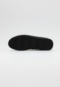 Černá gumová podrážka boty se zigzagovým vzorem a kulatým logem. Plný design, hladký povrch, minimalistický bez dalších detailů.