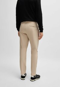 Pantaloni beige con una texture liscia, dotati di due tasche posteriori, dettaglio con logo e una vestibilità sagomata. Indossati con una maglietta nera a maniche lunghe e sneakers.