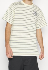 Randig t-shirt i ljusgul med marinblå horisontella linjer. Har en rund hals, korta ärmar och en cirkulär logotyp på bröstet. Mjuk tyg.