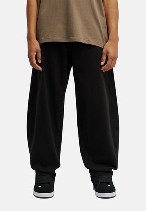 Persoon met losse zwarte broek, zwarte sneakers met witte zolen en een bruin topje, staand tegen een effen achtergrond.