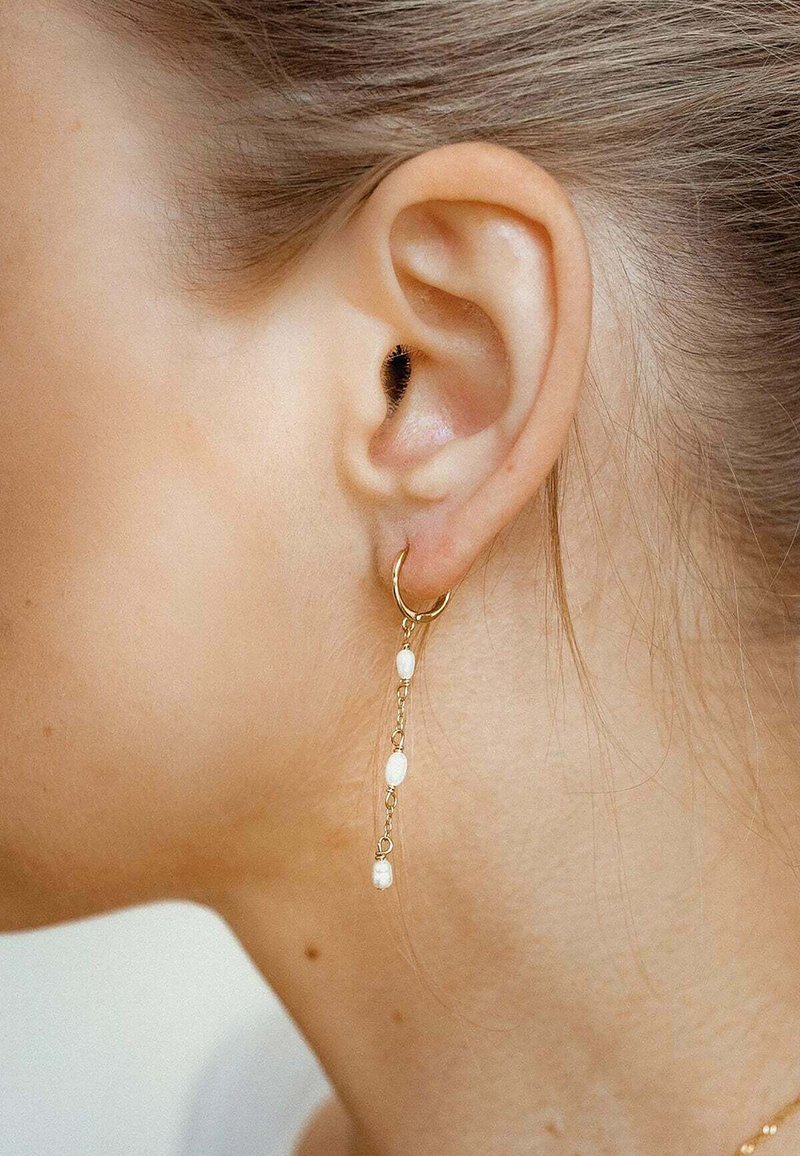 Pendiente de aro dorado con una delicada cadena que presenta pequeñas cuentas blancas redondeadas, colgando de la lóbulos de la oreja, mostrando un acabado pulido.