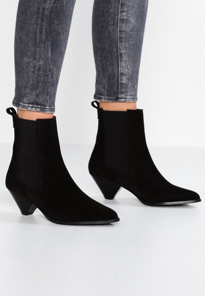 Botines de ante negros con punta afilada y tacón cónico. Cuentan con paneles laterales elásticos y una lengüeta para facilitar su uso.