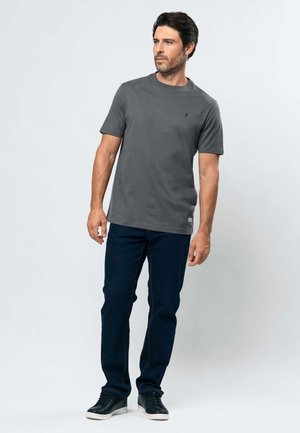 Moški s temnimi lasmi, oblečen v sivo kratko majico, temno modre kavbojke in črne superge, stoji pred preprostim svetlim ozadjem.