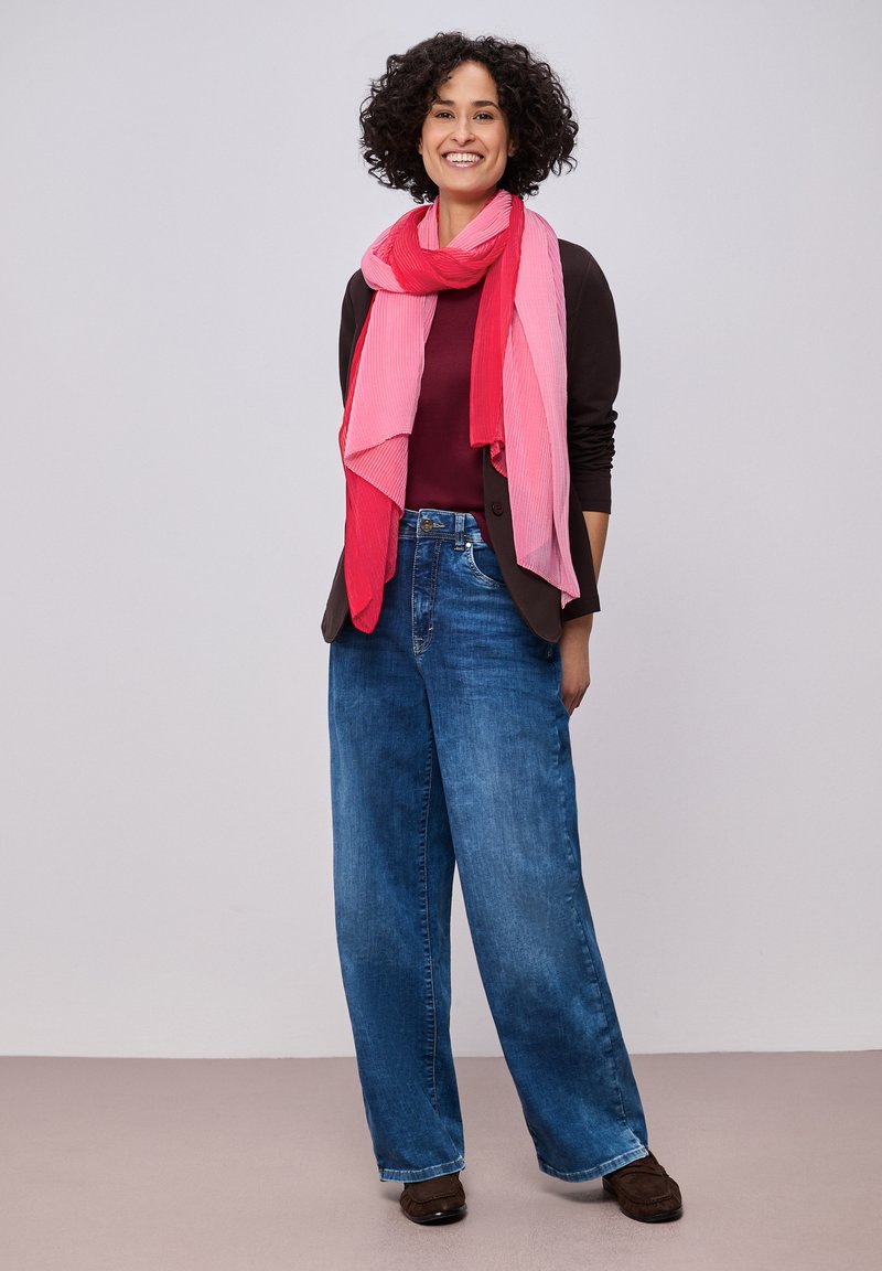 Femme souriante aux cheveux bouclés, portant une écharpe rouge et rose, un blazer foncé, un haut bordeaux, un jean bleu à jambes larges et des chaussures marron, debout contre un mur gris.