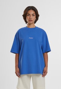 Blå oversized T-skjorte laget av bomull med rund hals, korte ermer og diskret "Forbudt" broderi på forsiden.
