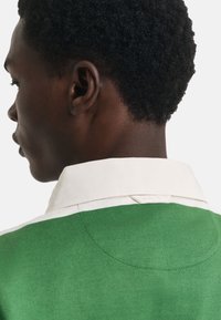 Grünes Strick-Poloshirt mit weißem Kragen, mit einem Etikett auf der Rückseite. Nahaufnahme des Ausschnitts und des Ohrenprofils.