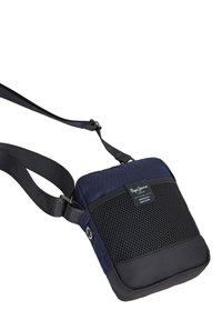 Jūras spēku un melns crossbody soma ar sieta tekstūru, ar rāvējslēdzēju, regulējamu siksnu un logo uzlīmi priekšpusē. Kompakta kvadrāta forma.