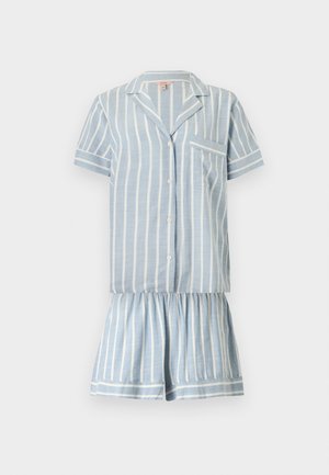 Jeune femme debout pieds nus portant une chemise à manches courtes boutonnée à rayures verticales bleu clair et blanc et un short assorti.