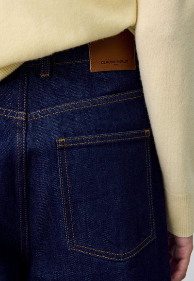 Jeans de mezclilla azul oscuro con un bolsillo trasero visible y una etiqueta de cuero marrón claro. Acompañados de un suéter de color crema.