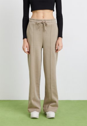 Beige Jogginghosen mit lockerer Passform, Kordelzugbund und vorderen Nähten. Aus weichem Material, kombiniert mit weißen Sneakers, getragen von einem Model.