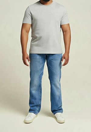 Mann steht und trägt ein hellgraues T-Shirt mit kurzen Ärmeln, blaue Jeans und weiße Turnschuhe vor einem einfarbigen beigen Hintergrund.