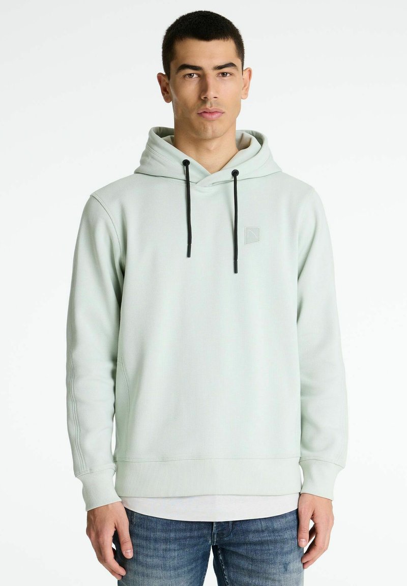 Sudadera verde claro con capucha ajustable, detalle de costura y un pequeño logo, combinada con jeans oscuros. Textura suave, diseño casual.