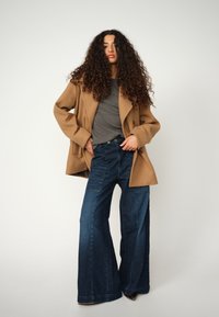 Young woman with long curly hair wearing a camel coat, gray top, and wide-leg dark blue jeans against a plain background.