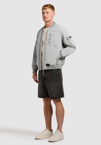 Jeune homme debout, portant une veste bomber gris clair, un t-shirt blanc, un short en denim noir et des baskets blanches sur un fond uni.