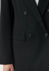 Blazer nero doppiopetto con quattro bottoni e tasca a pattina sul lato sinistro, indossato sopra una camicia bianca.