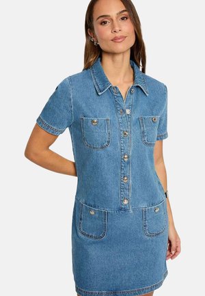 Femme portant une robe en denim bleu à manches courtes avec des boutons sur le devant et quatre poches frontales, debout devant un fond blanc.