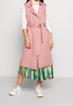 Femme portant un manteau rose sans manches ceinturé par-dessus un haut blanc à manches longues, une jupe verte fleurie et des baskets blanches, debout sur un fond uni.