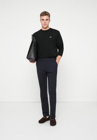 Zwarte shirt met lange mouwen en een klein logo, marineblauwe slim-fit broek en bruine instappers. Het model houdt een zwarte tote bag vast. Minimalistische achtergrond.