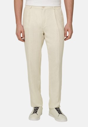 Man draagt beige op maat gemaakte broek en bijpassende sneakers met zwarte veters, staand tegen een effen achtergrond.