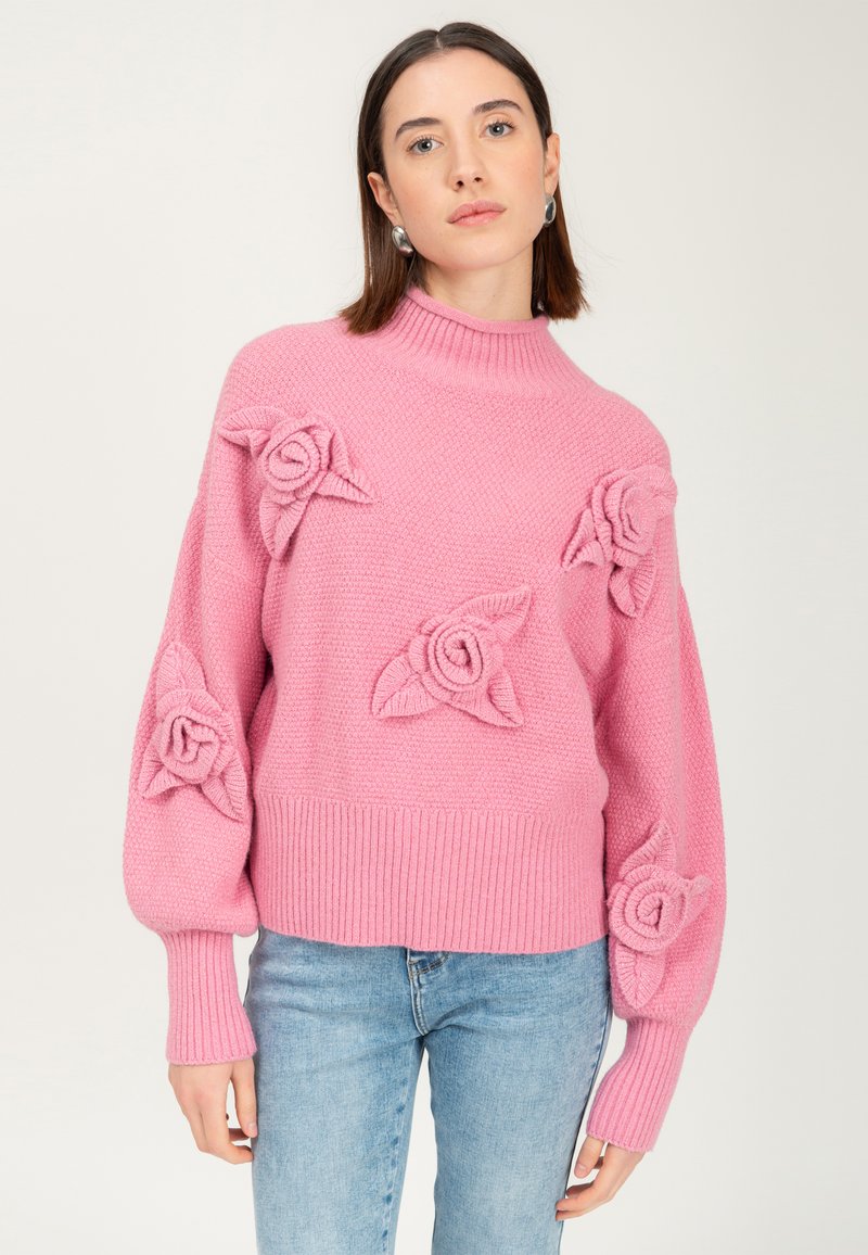 Donna che indossa un maglione a maglia rosa chiaro con motivi a rosa in rilievo e polsini a costine, abbinato a jeans blu chiaro, in piedi davanti a uno sfondo semplice.