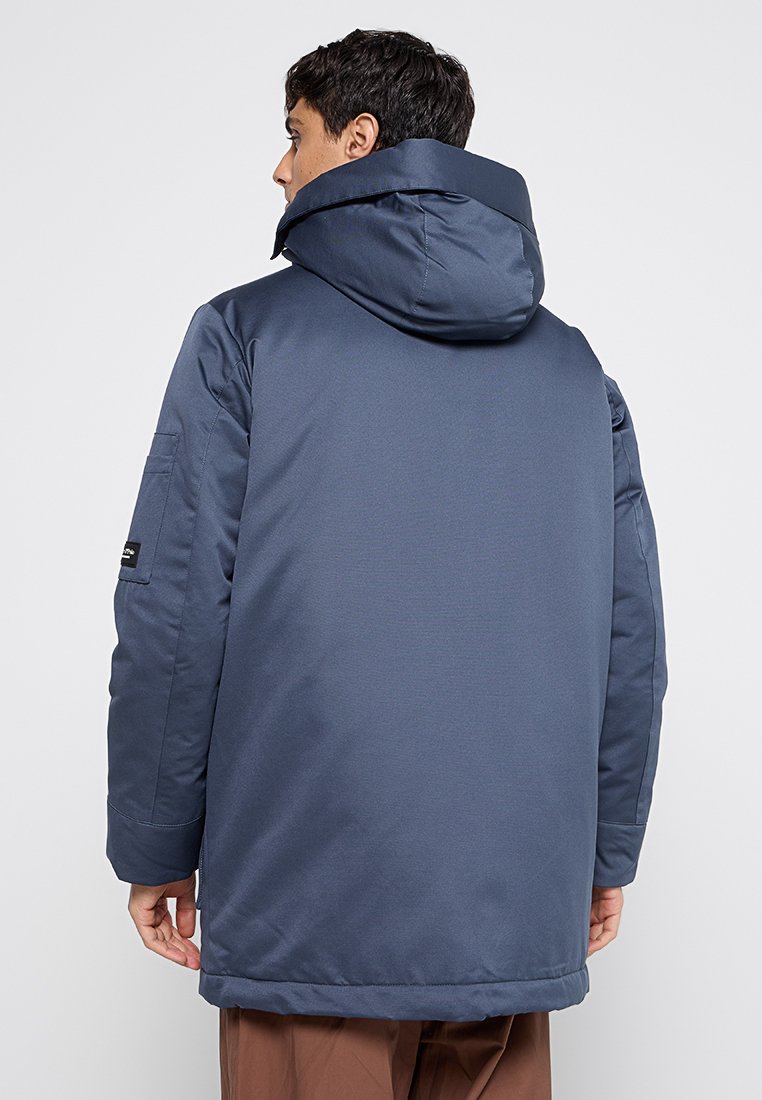 Marineblauwe waterdichte jas met een capuchon, met een ontspannen pasvorm, lange mouwen en een zak op de linkerarm. Glad textuur.