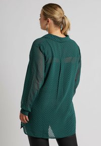 Turkusowa, przezroczysta bluzka w wąski wzór jodełki, z kołnierzykiem zapinanym na guziki i długimi rękawami; posiada rozcięcie u dołu i detal w postaci fałdy na plecach.