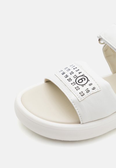 Vita sandaler med en bred rem som har tryckta siffror, mjukt material och rund tå. Tjock vit sula för komfort.