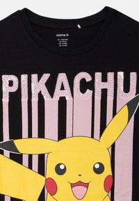 Melnā T-kreklā ir liela Pikachu grafika ar dzeltenām un melnām akcentiem. Virs tās ir teksts "PIKACHU" rozā pērlītēs, kas ir pretstatā vertikālām rozā svītrām.