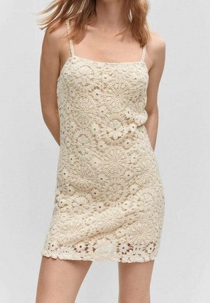 Femme portant une mini-robe en crochet sans manches de couleur crème avec des motifs floraux circulaires sur un fond gris uni.