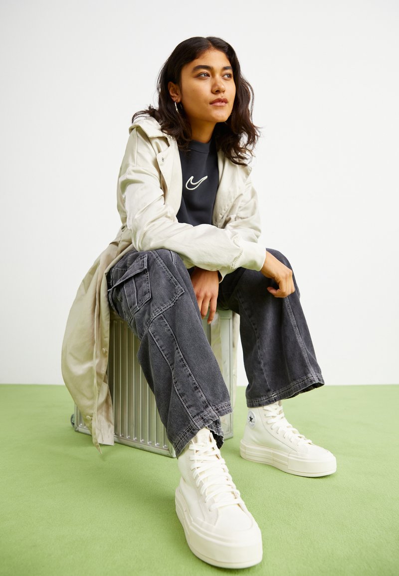 Cappotto lungo beige chiaro, felpa nera con logo, pantaloni cargo grigio scuro e sneakers bianche alte, seduto su una superficie metallica.