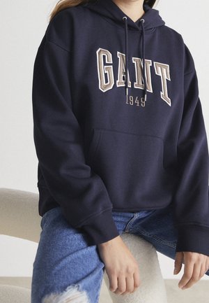 Person iført en mørkeblå GANT-hoodie med teksten "1949", kombineret med revnede blå jeans, siddende på en lysfarvet stol.