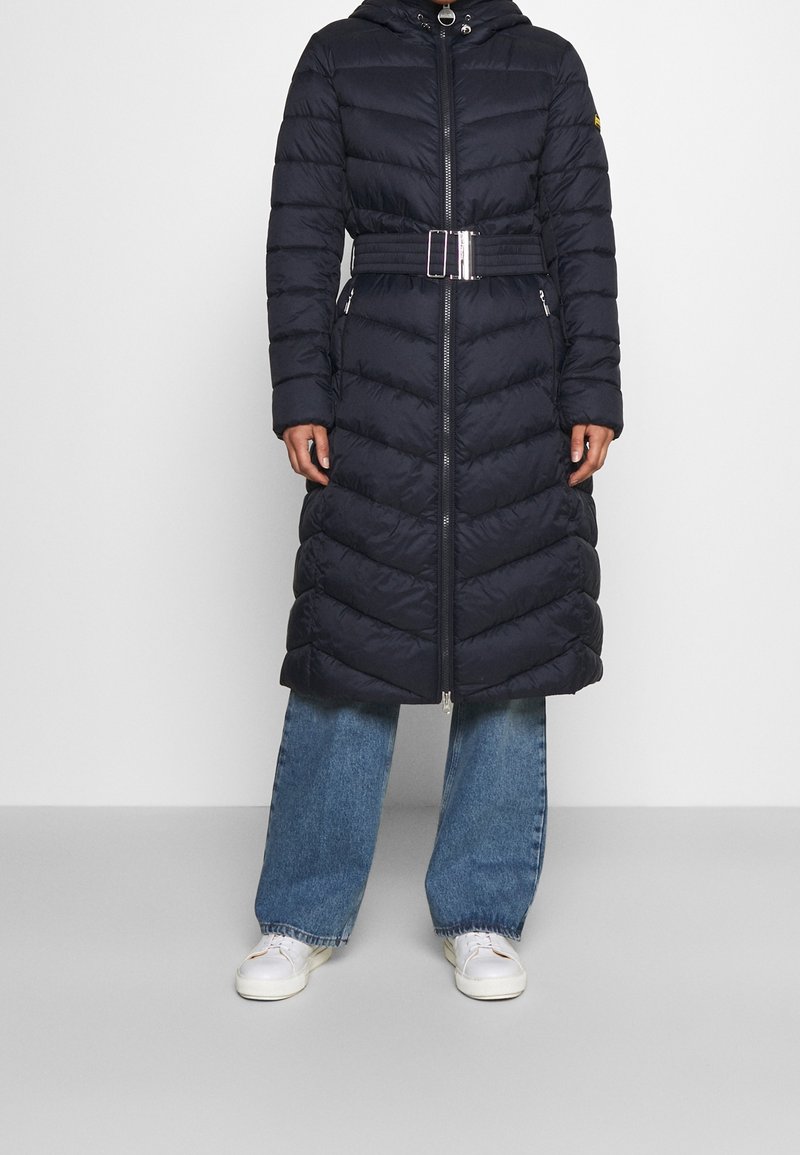 Langer marineblauer Stepp-Puffer-Mantel mit Kapuze, horizontalem Steppmuster, Reißverschluss und tailliertem Gürtel, kombiniert mit weitem Jeansschnitt und weißen Sneakern.