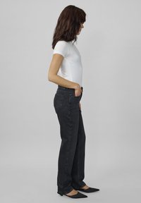 Svarta högmidjade jeans med rak passform, tillsammans med en åtsittande vit t-shirt. Modellen står på sidan mot en grå bakgrund.