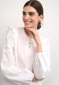 Blusa bianca con spalle arricciate, scollatura rotonda e tre bottoni in tonalità dorata. Maniche lunghe e svasate con polsini elasticizzati. Tessuto liscio.
