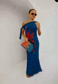Μπλε, off-shoulder φόρεμα με κόκκινο λουλουδάτο μοτίβο; Το μοντέλο κρατά μια υφασμάτινη μπλε τσάντα με σχέδιο πορτοκαλί ήλιο; καφέ σανδάλια.