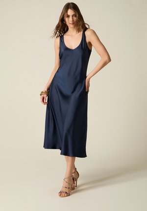 Kvinde iført en ærmeløs navy satin midi-kjole med en rund halsudskæring og højhælede sandaler med remme, stående mod en ensfarvet baggrund.